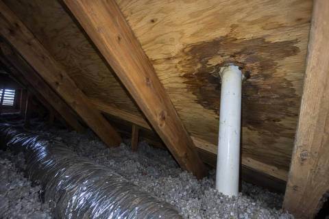 Underside of roof decking in an attic with a dark water stain around a white vent pipe, rafters, a silver duct and loose insulation below