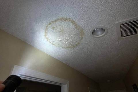 Brown ring-shaped water stain on a textured white ceiling beside a recessed light and an air vent, lit by a flashlight