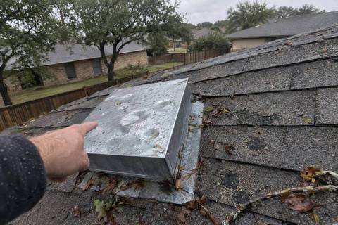 Square metal roof vent with round dents in its top beside shingles with dark bruised spots, wet leaves on the roof and a hand pointing at the vent