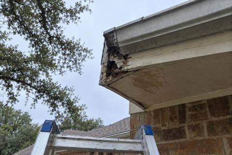 Corner of a roof edge seen from below with a rotted fascia board behind a gutter end and a stained, sagging soffit panel beside brick