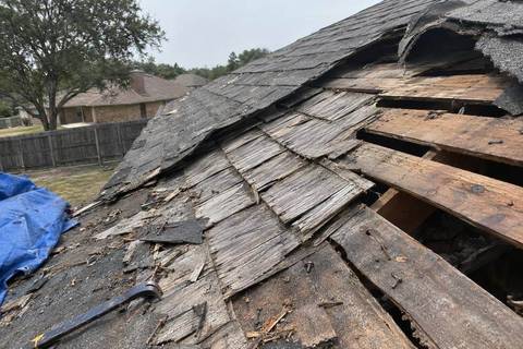 Asphalt shingles peeled back to show weathered gray wood shakes beneath, with spaced boards and gaps visible where the shakes are gone