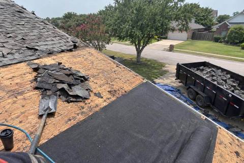 Roof seen from a ladder with one slope stripped to wood decking, felt rolled along the lower edge and a trailer of old shingles in the driveway