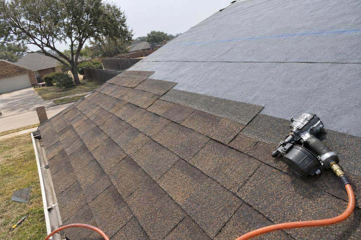 Roof slope partly covered in new brown dimensional shingles with gray underlayment above, a chalk line across it and a nail gun on an orange hose